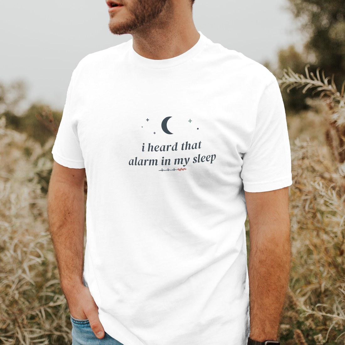 White tee reading “i heard that alarm in my sleep” in a clean angled studio shot